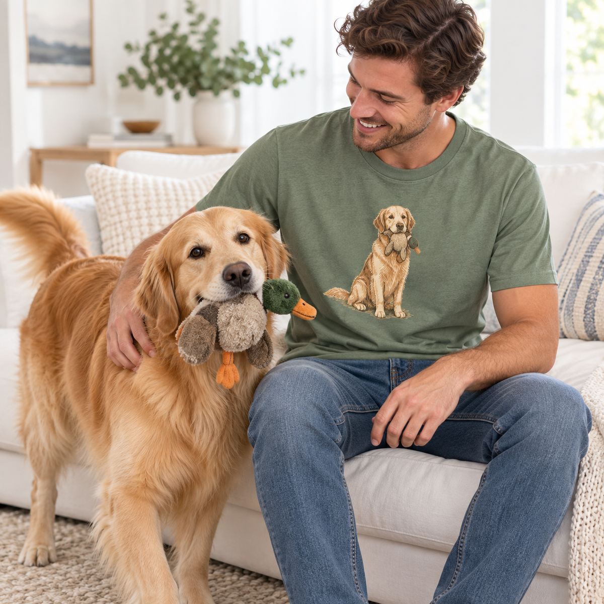 An adult wearing a sage Golden Retriever graphic tee sits with a Golden Retriever carrying a toy.